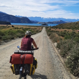 A woman riding a fully packed bicycle down a dirt road towards a lake, taken from the back
