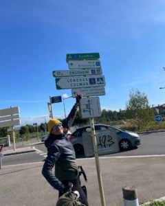 Man on a bicycle on a street corner pointing upwards towards street directional signs.