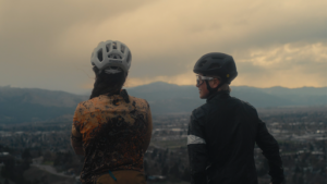 Two people wearing bicycle helmets standing at the top of a mountain looking at the valley